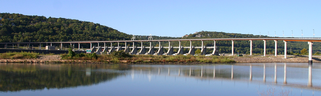 Long low bridge over a wide river during daytime, with green hills and clear blue sky in the background